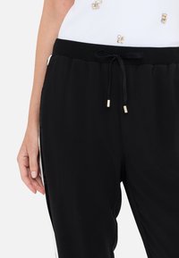 Primer plano de una persona que lleva pantalones negros con cordones dorados y una camiseta blanca con pequeñas decoraciones doradas.