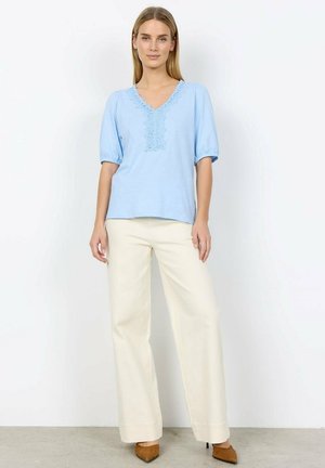 Vrouw staand, gekleed in een lichtblauw V-hals blouse met kantdetails, crèmekleurige wijde broekspijpen en bruine puntige hakken tegen een witte achtergrond.