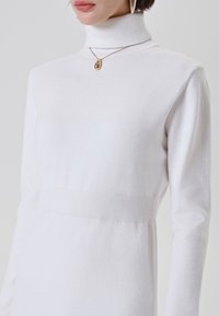 Donna che indossa un maglione bianco a collo alto e una collana con pendente dorato, ritagliata dalle spalle ai fianchi su uno sfondo semplice.