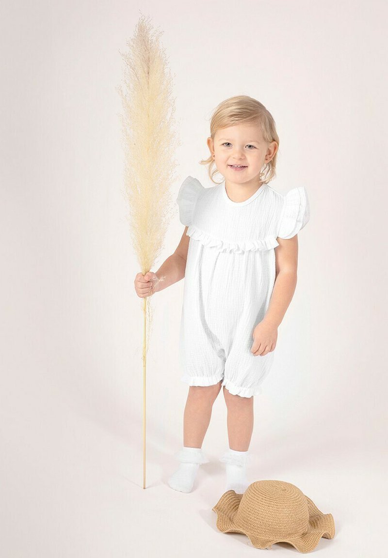 Biały teksturowany kombinezon z falbanowymi rękawami, stojące dziecko trzymające wysoki suchy łodygę trawy. Obok na ziemi leży słomkowy kapelusz.