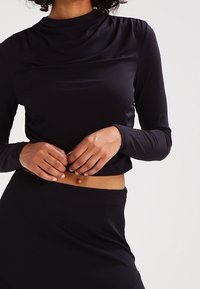 Svart långärmad topp med hög halsringning, figurnära passform och subtil draperingsdetalj. Tillverkad av ett mjukt, stretchigt tyg.