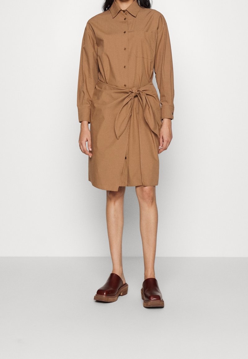 Abito camicia marrone a bottoni con maniche lunghe, dettaglio in vita annodato e colletto. Indossato con zoccoli marroni scuri e uno sfondo neutro.