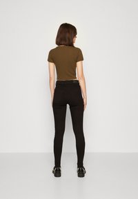 Olivi vihreä lyhyt t-paita yhdistettynä mustiin slim fit -farkkuihin. Näkyvä musta vyö, takataskut ja kiiltävät mustat kenkäyksityiskohdat. Neutraali tausta.