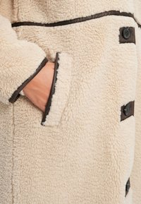 Κρεμ φλις παλτό με υφή, που διαθέτει σκούρα περιθώρια και τσέπη. Μαύρα κουμπιά προσθέτουν αντίθεση στο απαλά υφασμένο ύφασμα.