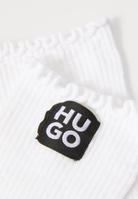 Hvide ribbede strømper med en flosset kant; har et sort logo-patch med "HUGO" i hvid tekst, der angiver mærkets identitet.