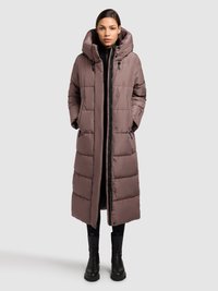 Ilgas rudos spalvos pūkinis paltas su nuimamu gaubtu, užtrauktuku, kišenėmis su užtrauktukais ir pūstu raštu. Dėvimas ant juodos palaidinės ir kelnių.