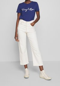 Modrá tričko s bílým logem "Tommy Hilfiger", bílé džíny s vysokým pasem a širokými nohavicemi, a béžové vysoké tenisky. Jednoduchý design a čisté barvy.