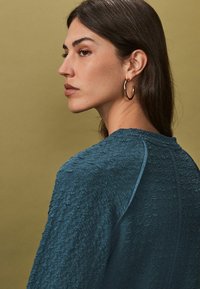 Teal textuurtrui met een ronde halslijn, schoudernaden en een losse pasvorm. Model draagt oorringen met lang, steil haar tegen een gouden achtergrond.