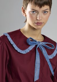 Blusa bordeaux con colletto volants blu chiaro e fiocco. Texture liscia, bottoni sul davanti e tessuto morbido. Ritratto in primo piano.