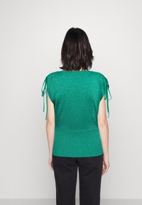 Groene, kortmouwen top met een gestructureerde, metallic afwerking. Beschikt over strikaccenten op de mouwen en een aansluitende zoom. G gedragen met zwarte broek.