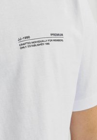 Balta medvilnės marškinėliai su juodu tekstu: "JJ-1990 PREMIUM" ir "KURIAMI INDIVIDUALIAI TIK NARIAMS. ĮSTEIGTA 1990."
