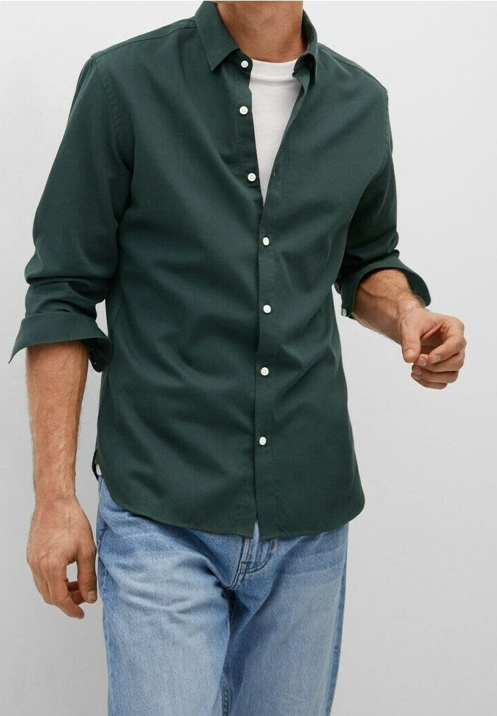 Homme portant une chemise boutonnée vert foncé par-dessus un t-shirt blanc avec manches retroussées et un jean bleu clair sur fond uni.