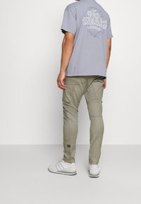 Grå t-shirt med texten "Nej tack" på baksidan, parat med olivgröna cargobyxor som har flera fickor och en texturerad yta. Vita sneakers.