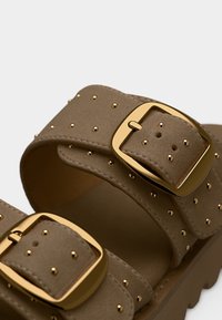 Gros plan sur des lanières de sandales marron ornées de clous dorés et de boucles carrées dorées sur une semelle texturée.