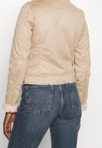 Chaqueta de ante sintético beige con diseño acolchado, acentos de puños de borreguito y un corte corto. Combinada con unos vaqueros de denim azul.