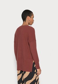Sweater tricotado marrom com textura canelada, ombros caídos e fendas laterais. Usado com calças largas estampadas em bege e preto.