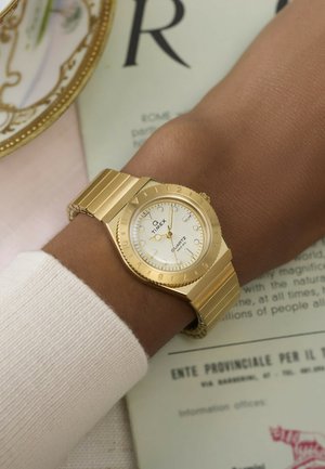 Orologio da polso Timex color oro con quadrante bianco e cinturino in metallo su un polso dalla pelle scura sopra un maglione chiaro, appoggiato su un documento.