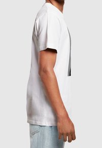 Bílá bavlněná tričko s krátkými rukávy a kulatým výstřihem, s grafickým designem na přední straně, zobrazené z boku. Světle modré džíny.