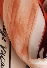 Gros plan d’un tissu avec des motifs de coups de pinceau orange et beige couvrant partiellement un texte en cursive noire sur un fond clair.