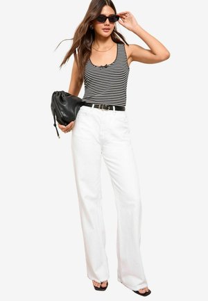 Schwarz-weiß gestreiftes Tanktop mit einer Schleifenakzent, kombiniert mit hochgeschnittenen weißen weiten Jeans und schwarzen Accessoires, darunter Sonnenbrille und Tasche.