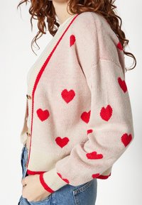 Cardigan rosa a maglia con motivi a cuore rossi, caratterizzato da un collo a V, polsini a coste e una vita coordinata. Presenta bottoni dorati sul davanti.