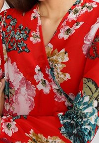 Blusa avvolgente rossa a fiori con maniche lunghe, caratterizzata da un mix di fiori rosa, bianchi e verde teal. Realizzata in tessuto leggero e morbido con scollatura a V.