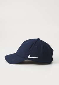 Nike-keps i marinblått tyg, med en vit swoosh-logotyp framtill, rundad skärm och ventilationshål på sidorna.