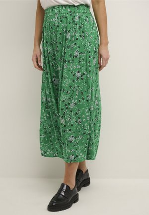 Kvinde iført grøn blomsterprintet midinederdel med opsamlet talje og sorte kraftige læderloafers mod en ensfarvet baggrund.