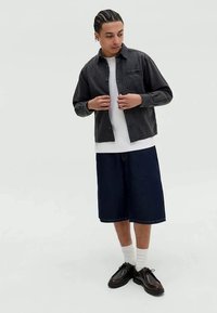 Zkontrolovaná černá košile bunda přes bílé tričko, kombinovaná s tmavými džínovými šortkami a černými šněrovacími botami s viditelným šitím a kapsami.