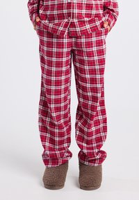 Rode en witte geruite pyjamabroek gedragen met een bijpassende top, handen in de zakken, en bruine fuzzy slippers op een witte achtergrond.