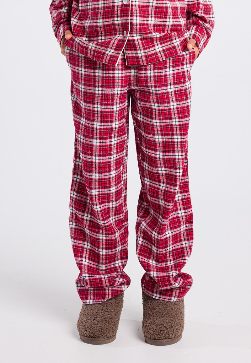 Rode en witte geruite pyjamabroek gedragen met een bijpassende top, handen in de zakken, en bruine fuzzy slippers op een witte achtergrond.