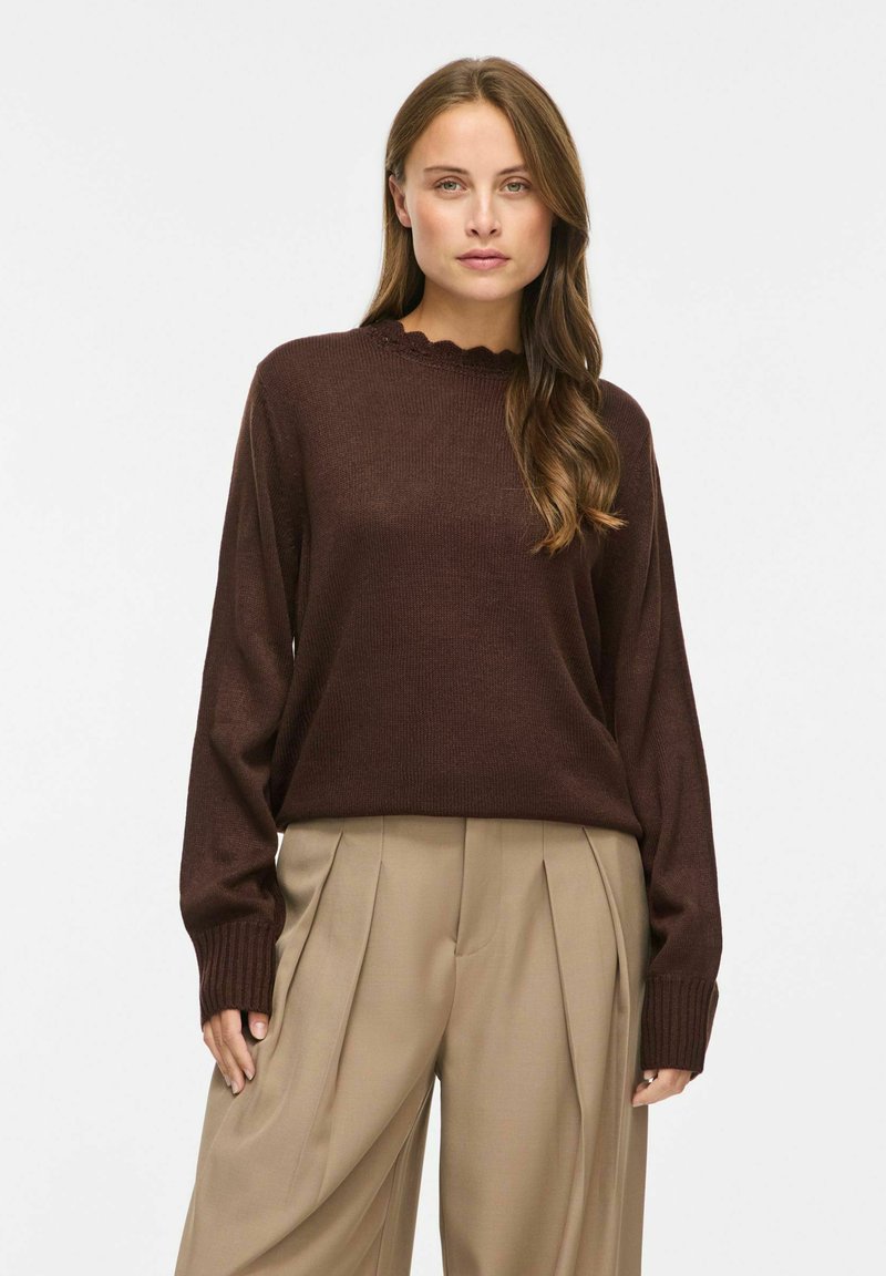 Femme aux longs cheveux bruns portant un pull marron foncé et un pantalon ample beige plissé, debout devant un fond blanc uni.