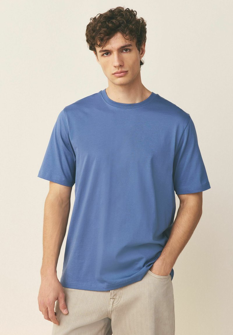 Junger Mann mit lockigem Haar, der ein schlichtes blaues T-Shirt mit kurzen Ärmeln und hellbeige Hose trägt, steht mit einer Hand in der Tasche.