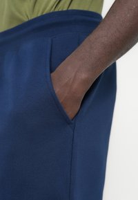 Main à la peau foncée insérée dans la poche d’un pantalon bleu marine, vue partielle d'une chemise verte au-dessus de la ceinture.