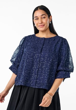 Mörkblå blus med en texturerad, broderad design som har paljetter. Korta, puffiga ärmar och rund halsringning; knäppning framtill.