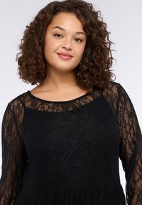 Czarna koronkowa sukienka z długimi rękawami, okrągłym dekoltem i falującym dołem. Charakteryzuje się misternymi wzorami kwiatowymi i teksturowaną tkaniną.