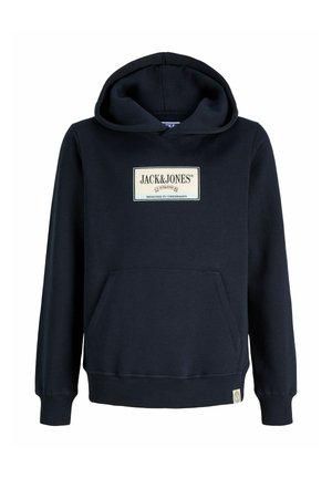 Garçon debout avec un sweat à capuche noir Jack & Jones, un jean bleu ample et des baskets blanches sur un fond clair uni.