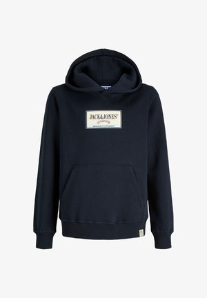 Chłopiec stojący w czarnej bluzie z kapturem Jack & Jones, luźnych niebieskich jeansach i białych trampkach na jednolitym jasnym tle.