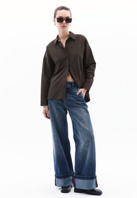 Camicia marrone a bottoni, jeans blu di jeans a gamba larga con risvolto, scarpe nere e occhiali da sole. Design semplice e strutturato, vestibilità casual.