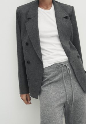 Femme portant un blazer croisé gris foncé, un t-shirt blanc et un pantalon gris clair à cordon, devant un fond clair uni.