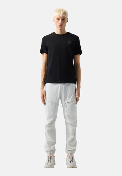 Ung person med kort blondt hår iført en sort T-shirt, lysegrå cargobukser og hvide sneakers mod en ensfarvet baggrund.