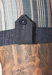 Gestreiftes Stoffhemd mit blauen und orangefarbenen Streifen, Denim- Akzenten und einer geknöpften Klappe. Sichtbares Tattoo auf der Haut unter dem Hemd.