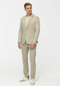 Beige textúrájú öltönyt viselő férfi, akihez illik a bézs színű nadrág és ing, valamint fehér sportcipő. A háttér sima fehér.