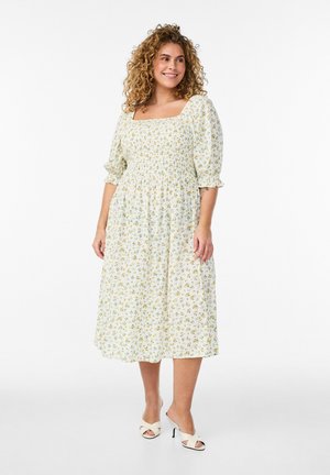 Kvinde med krøllet hår iført en hvid midikjole med gult blomsterprint, puffede ærmer og hvide hælsandaler, smilende og kigger mod højre.