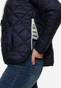 Marinblå kviltad jacka med sidoredskap och vita kontraster. Blå jeans med framficka. Texturerat material; lätt glans på ytan.