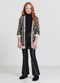 Long cardigan en motif léopard crème et noir, associé à un pantalon noir ajusté et des chaussures noires. Texture douce, coupe ample et design ouvert.