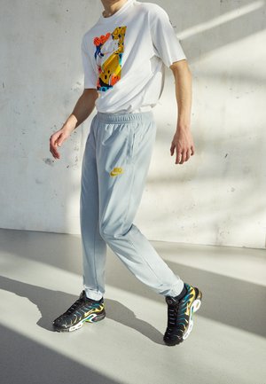 Osoba ubrana w białą koszulkę z nadrukiem, jasnoszare spodnie dresowe Nike oraz czarne buty sportowe z niebieskimi i żółtymi akcentami, chodząca po gładkiej podłodze wewnątrz budynku.