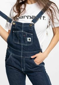 Dunkelblaue Denim-Latzhose mit verstellbaren Trägern, frontalen Taschen und einem kleinen Markenlogo-Patch. Kombiniert mit einem weißen Logo-T-Shirt.