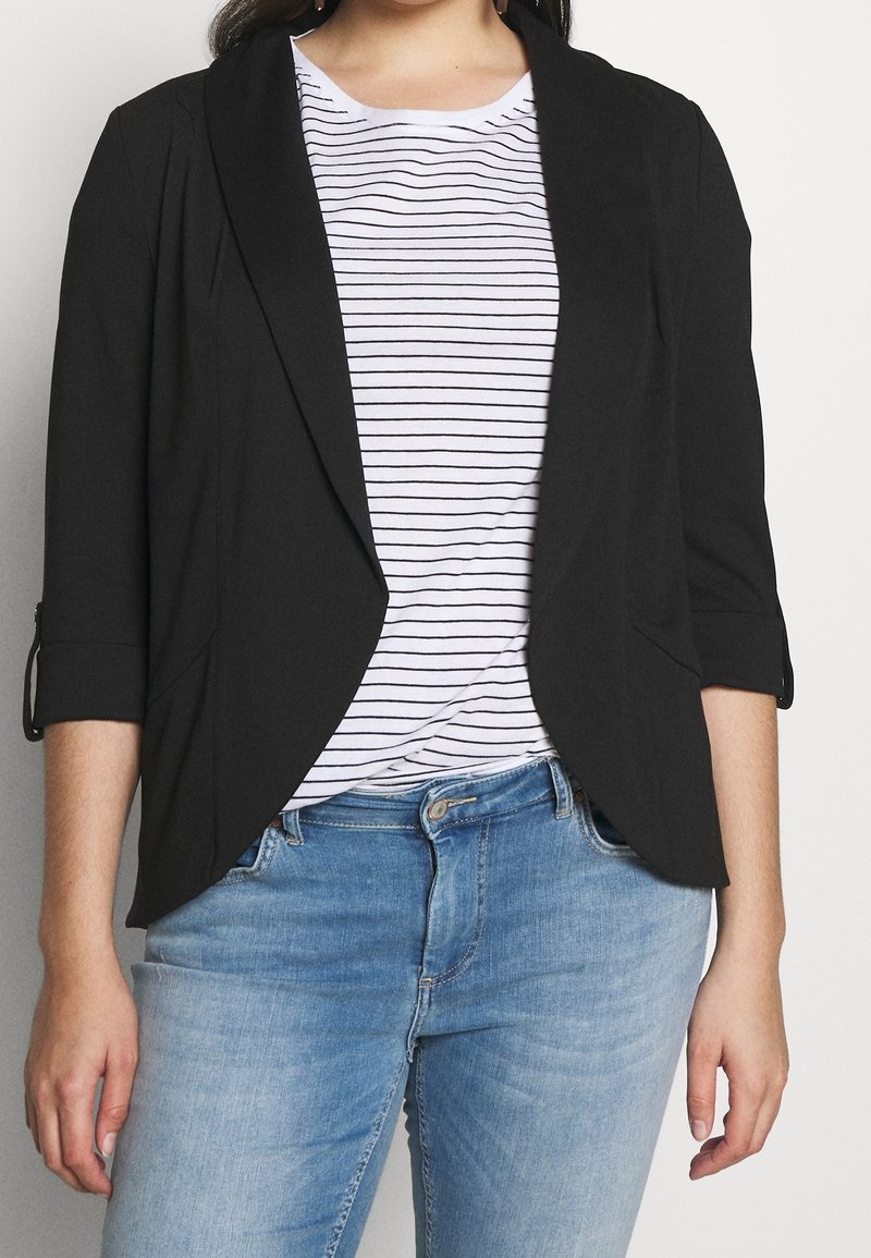 Blazer noir avec un col châle, des manches trois-quarts et des lignes épurées, porté sur une chemise rayée et un jean bleu clair. Texture de tissu lisse.