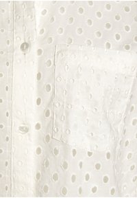 Camicia bianca con bottoni, caratterizzata da un tessuto testurizzato con ritagli circolari, una singola tasca e una chiusura con bottone in plastica trasparente.